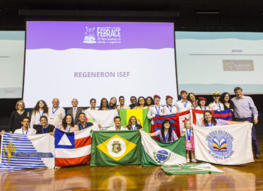 Grupo grande de estudantes e professores posando em um palco durante um evento científico. Ao fundo, um telão exibe “FEBRACE – 24ª feira brasileira de ciências e engenharia” e “Regeneron ISEF”. Os participantes, de diferentes idades e etnias, usam crachás e medalhas e seguram diversas bandeiras e estandartes de escolas e estados brasileiros. Alguns vestem uniformes escolares, incluindo alunos com boinas vermelhas. O clima é de celebração e conquista, com todos sorrindo para a foto.
